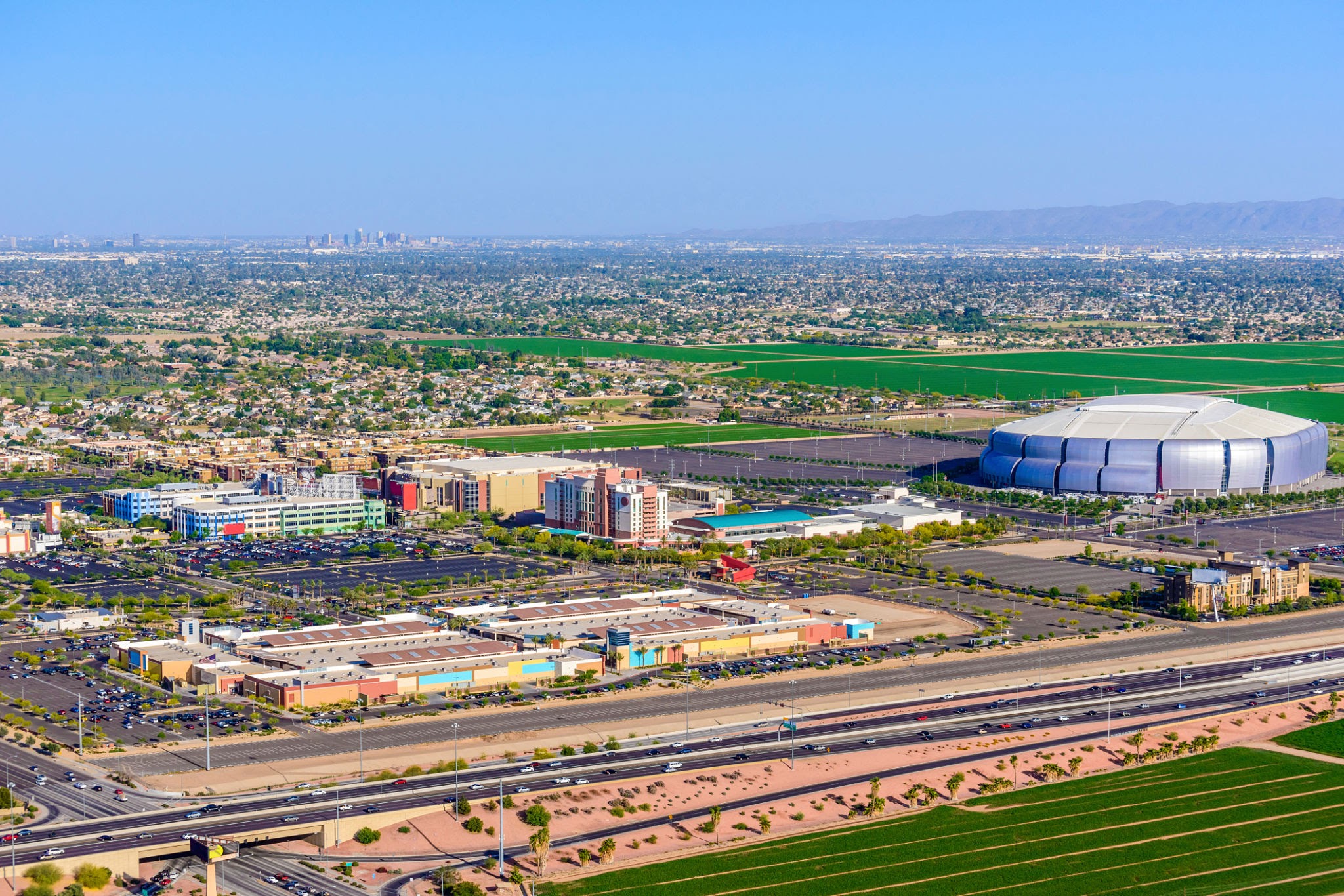 An outlet mall, hotels, and various other shopping and entertainment facilities in Glendale Arizona (a suburb of Phoenix, Arizona). Phoenix skyline is visible in the distance on the horizon. The University of Phoenix Stadium is home to the 2015 championship game XLIX.