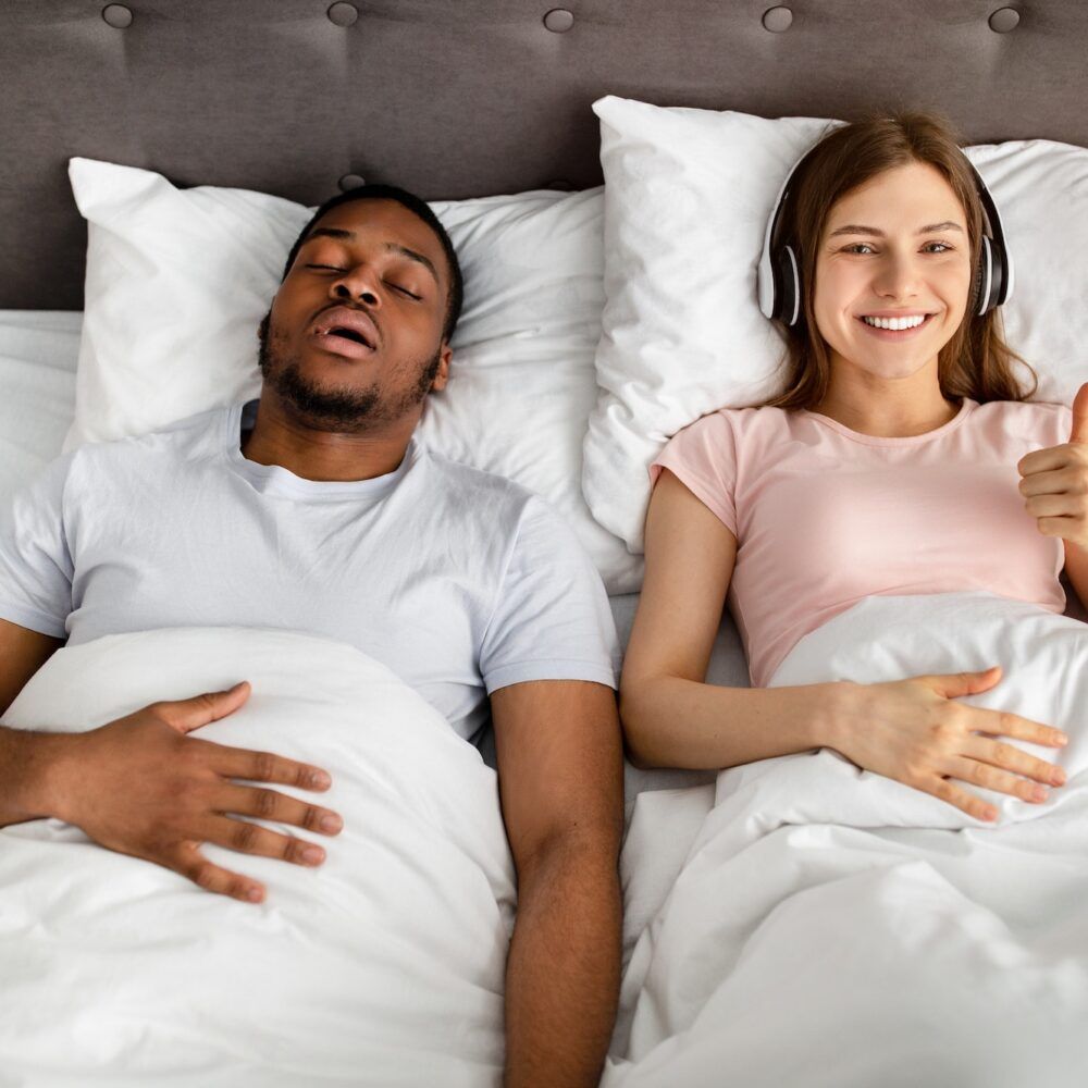 Happy young woman wearing headphones and showing thumb up gesture while her husband snoring on bed