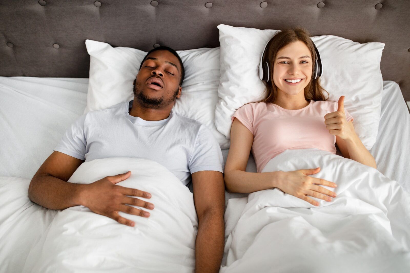 Happy young woman wearing headphones and showing thumb up gesture while her husband snoring on bed