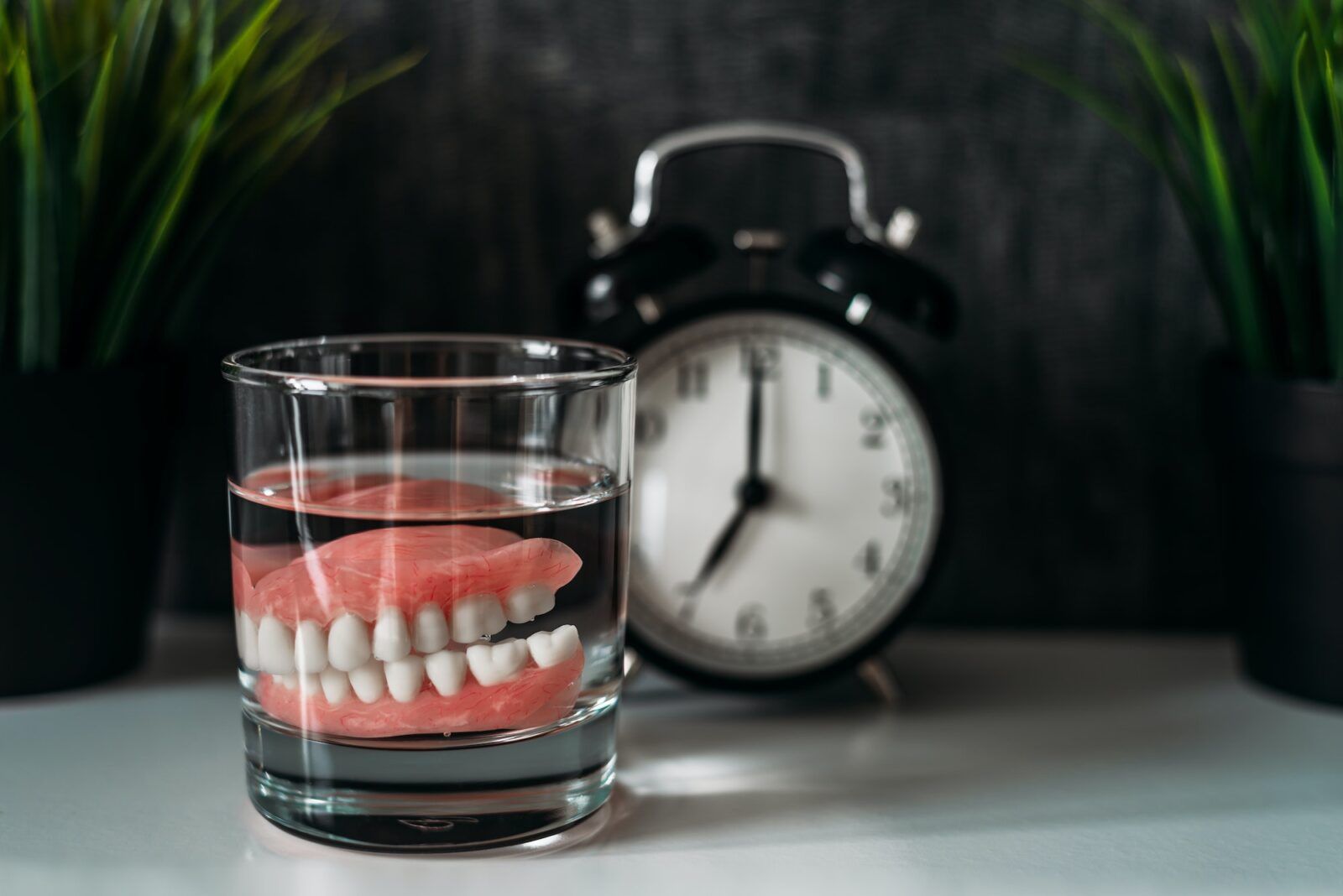 An acrylic denture in a glass of water and a clock in the background. Dentures or false teeth