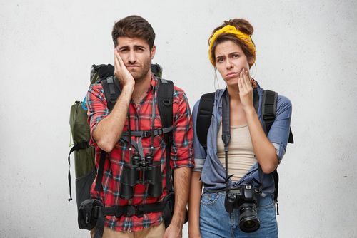Studio shot of stylish young male and female hipster tourists or hitchhikers holding hands on their cheeks suffering from toothache, looking in camera with painful expressions while traveling together